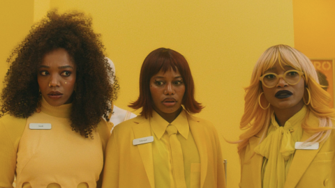 Three women with name tags, styled in yellow outfits and bold hairstyles, standing against a yellow background.