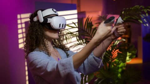 Woman wearing a VR headset and holding motion controllers in a colourful, plant-filled room.