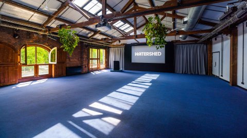 Open heritage event space with exposed timber beams, skylights, hanging plants, and a screen displaying “WATERSHED.”