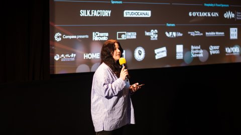 A woman speaks into a microphone on stage in front of a cinema screen displaying sponsor and partner logos
