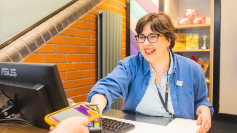 A Box Office assistant smiles as they present a card reader to a customer for payment