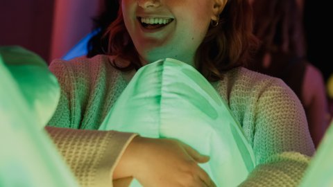 A young white woman is laughing whilst cuddling a big inflatable lit up robot.