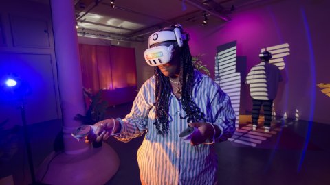 A young Black woman stands in a VR headset holding onto two controllers, a young Black man stands in the background. The room is pink and blue.