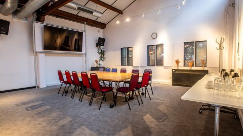 Conference room with a large table surrounded by red chairs, a TV, clock, and decorative plants in a well-lit space.