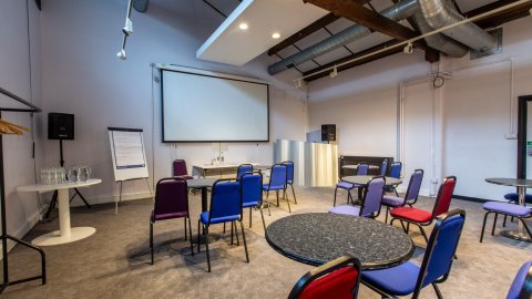 Modern meeting room with round tables, colorful chairs, a large screen, and audio equipment, designed for presentations or discussions.