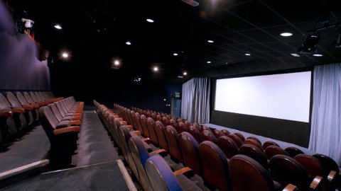 A dimly lit cinema hall featuring rows of red upholstered seats facing a large white screen, with curtains on one side.