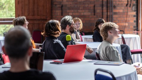 A group of people sitting at tables in a venue, with a vibrant red laptop and a green microphone in the foreground.