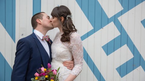 A couple shares a kiss, dressed elegantly, with the bride holding a colorful bouquet against a patterned blue backdrop.
