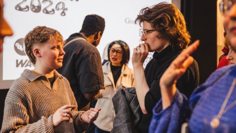 A group of young adults engaged in conversation at an event, with a backdrop displaying "Inside Screen 2024." Casual attire and animated expressions.