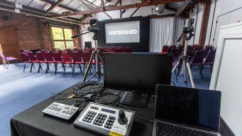 A room filled with red chairs, a projection screen showing "WATERSHED," and video equipment set up on a black table.