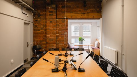 A modern meeting room featuring a wooden table with microphones, brick walls, and a bright window nook with plants.