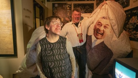 Andrew and Eden Kotting smile in a dimly lit room. Behind them, a large piece of fabric features a printed image of a person in a suit. A sense of joy is conveyed.
