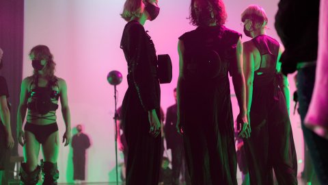 People in dark, avant-garde attire and masks stand under dramatic pink and green lighting on a stage, creating a mysterious, edgy atmosphere.