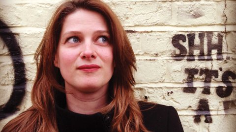 Person with long reddish hair and black coat standing against a white brick wall with graffiti reading "SHH IT'S A".