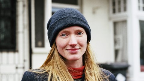 Person with long blond hair wearing a navy blue beanie and coat standing outdoors in front of a building.