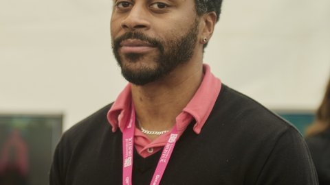 A Black man with a beard stands indoors wearing a black sweater and a red collared shirt. He has a thoughtful expression and wears a pink lanyard.