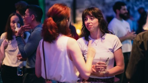 Two women chatting at a dimly lit social gathering, one holding a drink. Other people are blurred in the background.