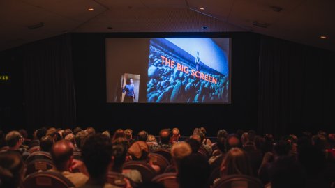 Audience watches a large cinema screen displaying “THE BIG SCREEN” in a dark theater.