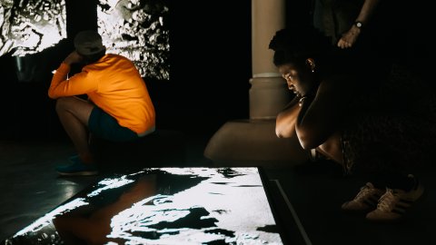 Two people sit in a dim room, illuminated by a bright projected image on the floor.