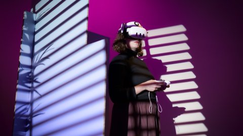 Person wearing a white VR headset and holding controllers, illuminated by striped window shadows against a purple wall.