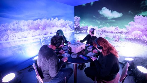 Four people wearing headphones interact with a projector and materials at a table in an immersive blue-purple landscape installation.