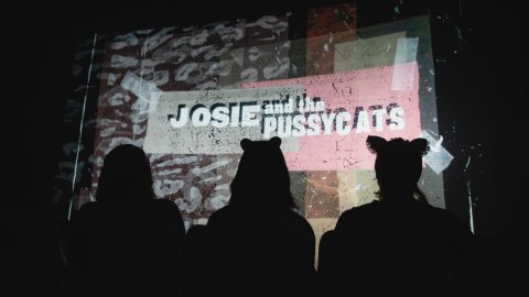Three silhouetted viewers watch a projected graphic reading “Josie and the Pussycats.”