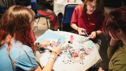 Two people craft a collage with scissors and magazines at a busy table.