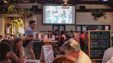 Bustling Watershed Cafe & Bar with diners at tables, a chalkboard menu, hanging plants, and a projection screen.