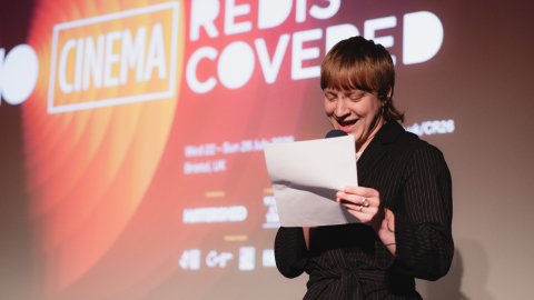 A young person in front of a screen displaying a Cinema Rediscovered slide speaks to an audience from their notes