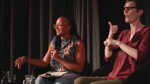 A woman speaks into a microphone while a sign language interpreter gestures beside her on a dimly lit stage.