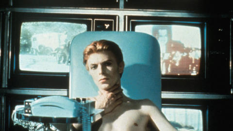 David Bowie sitting in a chair in front of TV screens