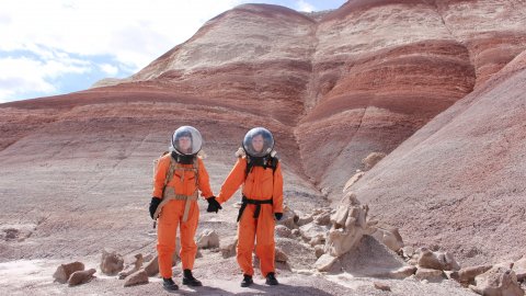 Two women in spacesuits in a barren landscape holding hands