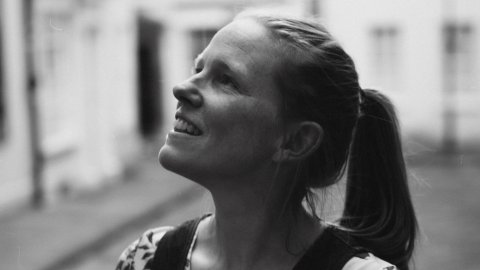 Photo of Kate Arthurs - a smiling woman with tied back light coloured hair, standing in a street, is looking up at her surroundings