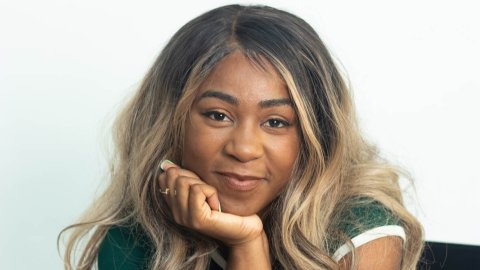 A young Black woman with long blonde hair rests her head on her chin and smiles softly into the camera.