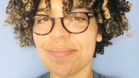A person with curly, multi-tonal hair poses against a soft blue background, wearing a green shirt.