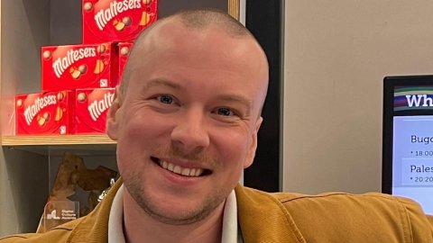 A person in a brown jacket stands casually, with Maltesers boxes visible in the background and a screen displaying event information nearby.