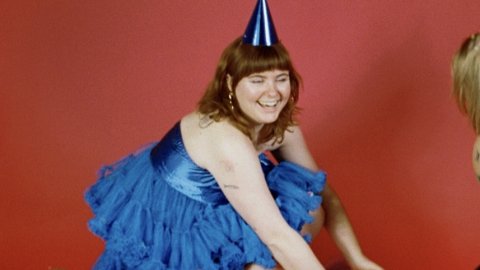 A person in a blue dress and party hat kneels on a red background, reaching for colourful balloons and surrounded by confetti.