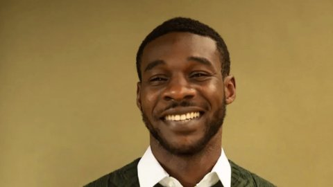 A person wearing a green cable-knit sweater over a white collared shirt stands against a neutral background.