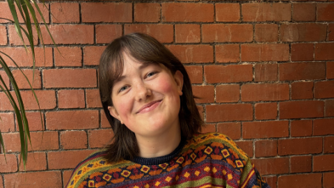 Person in a colorful patterned sweater and light blue jeans sitting on a black chair at a wooden table, with a brick wall background.