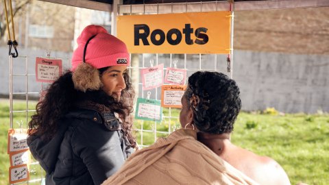 Play:Disrupt Creative High Streets Project, two people outside looking at note cards under an orange sign saying 'roots'