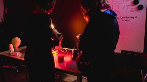 Two people examine a small object at a cluttered worktable under red light, beside a whiteboard covered in sketches and notes; the mood is focused and tense.