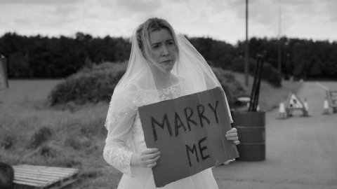 A bride wearing a veil holds a cardboard sign that reads "MARRY ME," standing by a roadside with a mailbox nearby.