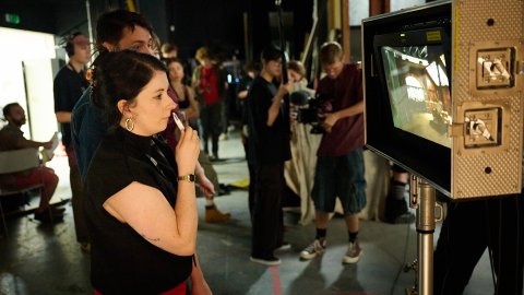 A woman attentively observes a monitor on a busy film set, surrounded by crew members and equipment. The scene conveys focus and collaboration.