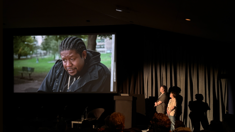 Audience watches a film screening in a dark theater while two presenters stand near a podium beside the screen.