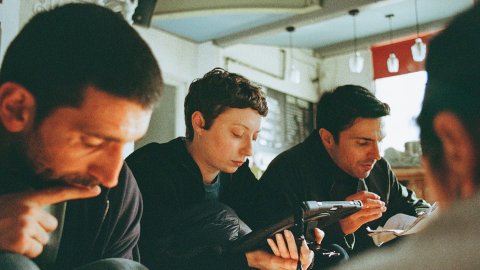 Behind the scenes on the shoot of Surviving Earth. Three people sit closely in a cozy café, a women looks at a handheld screen. The background shows warm lighting and a relaxed atmosphere.