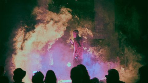 Performer holding a microphone on a foggy, dimly lit stage with an audience silhouette in the foreground.