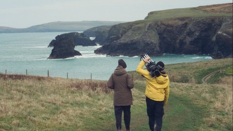 Two people walk along a grassy coastal path with cliffs and ocean in the background, one carrying a camera tripod.