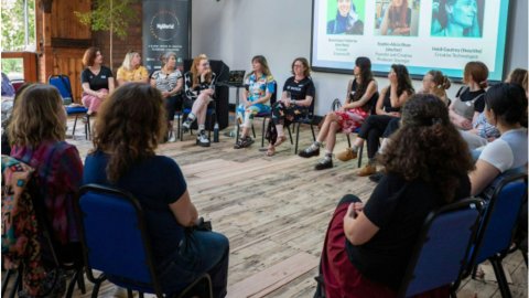 Diverse audience listens to women speakers seated in a circle during a community presentation, with portraits and speaker names projected behind them.