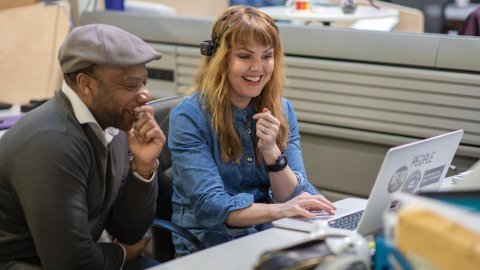Two coworkers smile while reviewing work on a laptop at an office desk; a collaborative, friendly atmosphere fills the workspace.