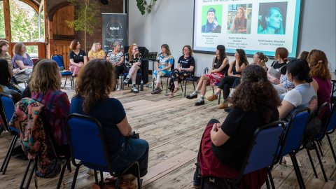 Diverse audience listens to women speakers seated in a circle during a community presentation, with portraits and speaker names projected behind them.